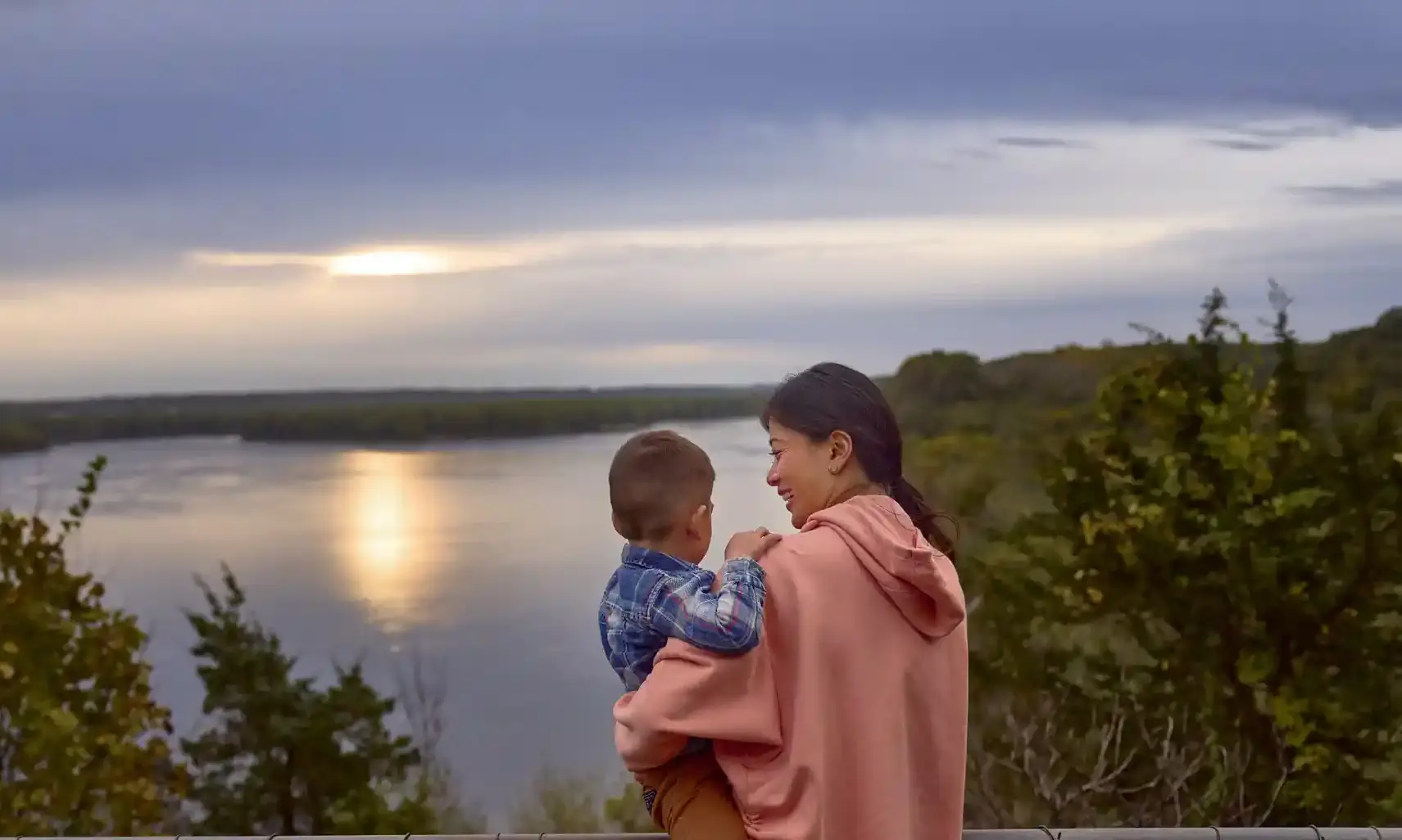 Mother and child standing on a hill overlooking a lake at sunset.